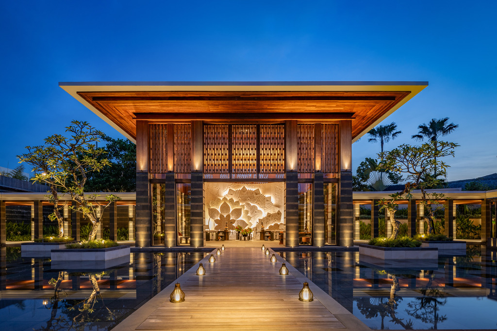 Regent Bali Canggu a dramatic dusk arrival pavilion with a timber-louvred façade, a glowing sculptural floral installation behind, a lantern-lined timber boardwalk, and flanking reflecting pools.