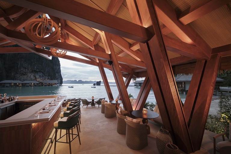 Lagen Island Resort El Nido bar and dining space with dramatic exposed dark timber A-frame roof structure, high bar stools along a counter and woven rattan low seating overlooking the bay at dusk.