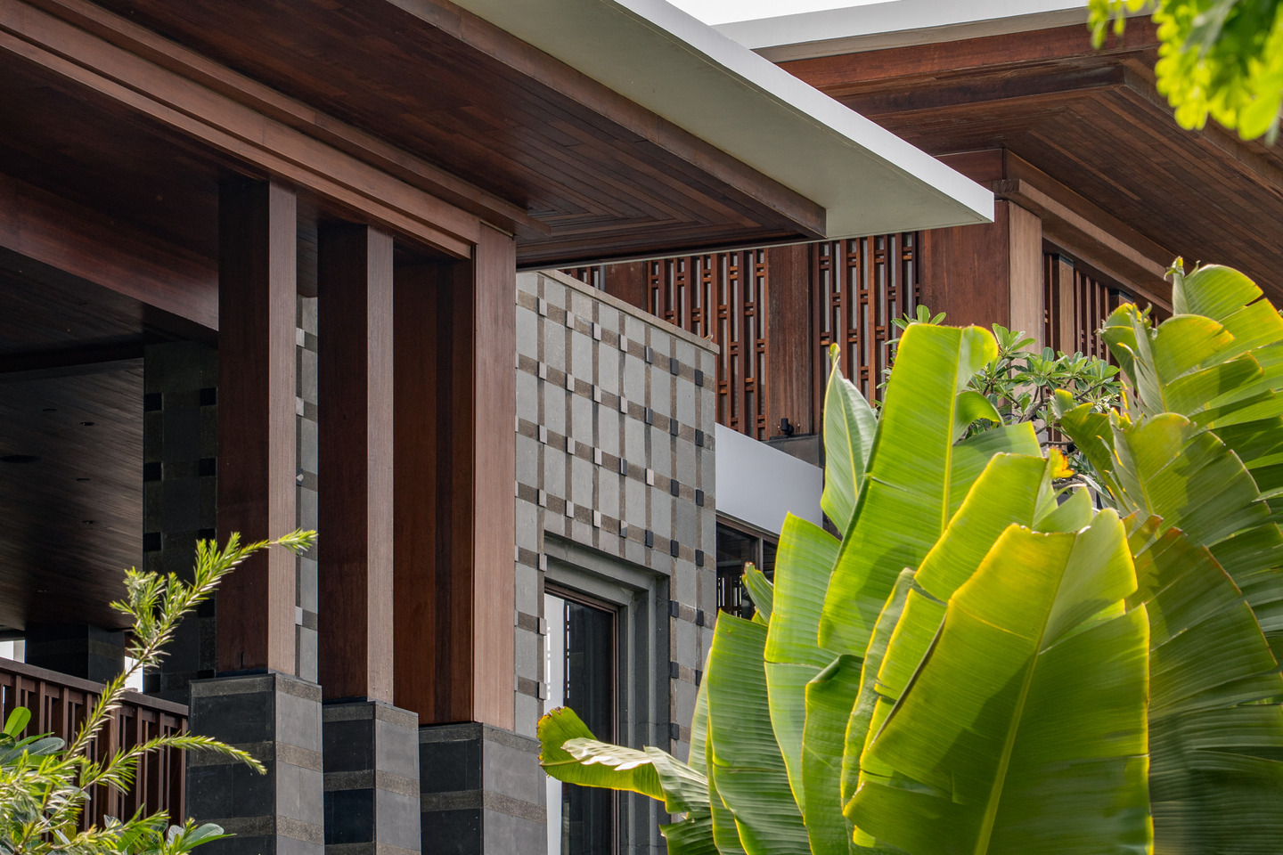 Regent Bali Canggu a close-up of the resort's layered façade detailing showing dark timber eaves, louvred balustrades, a woven stone cladding pattern, and lush banana and tropical foliage in the foreground.